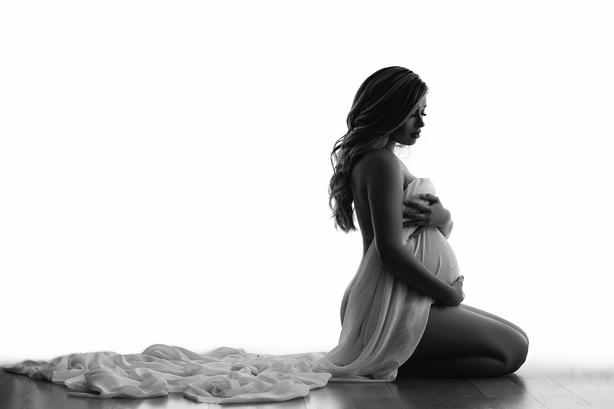 mom to be kneels on the floor while covering her bump with long white sheets Atlanta gynecology