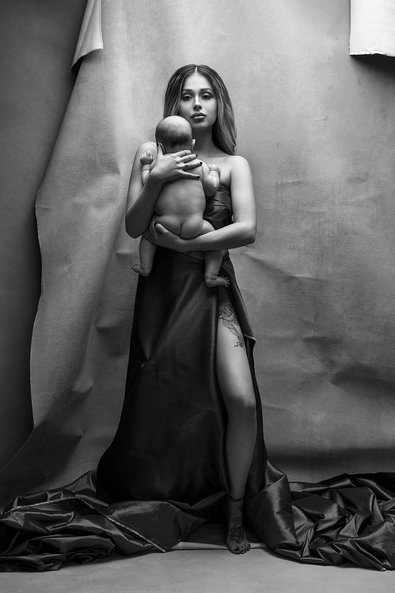 Woman stands in a studio holding her naked newborn wearing a long gown