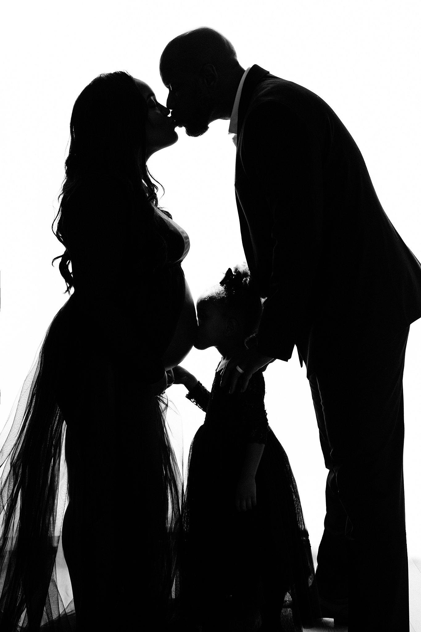 silhouette of a mom to be and dad kissing while their daughter kisses her bump