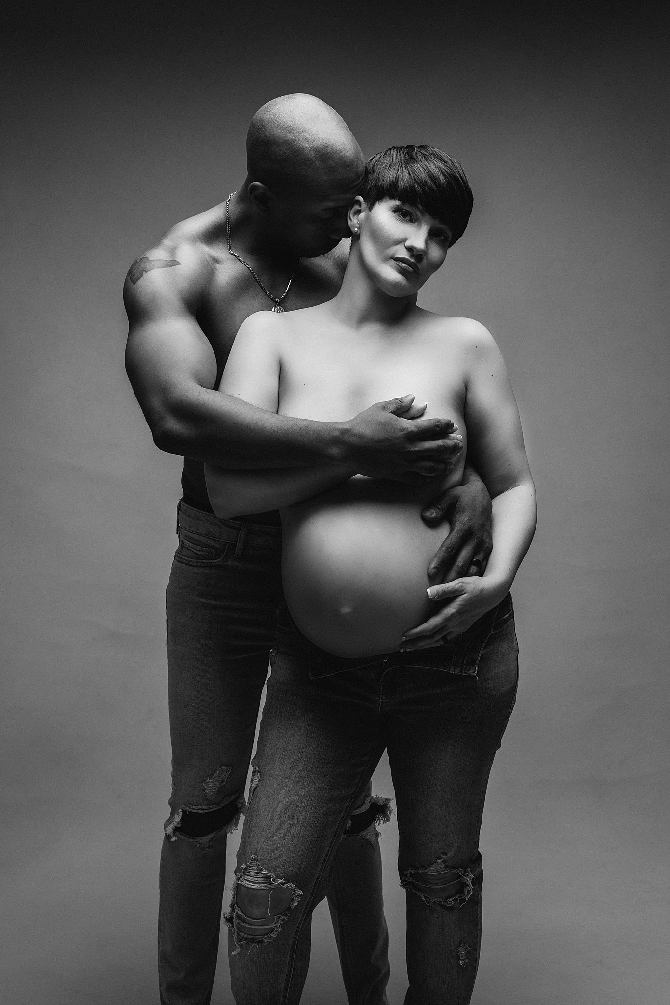 Pregnant woman wears ripped jeans in a studio being hugged from behind by man in jeans athens midwives