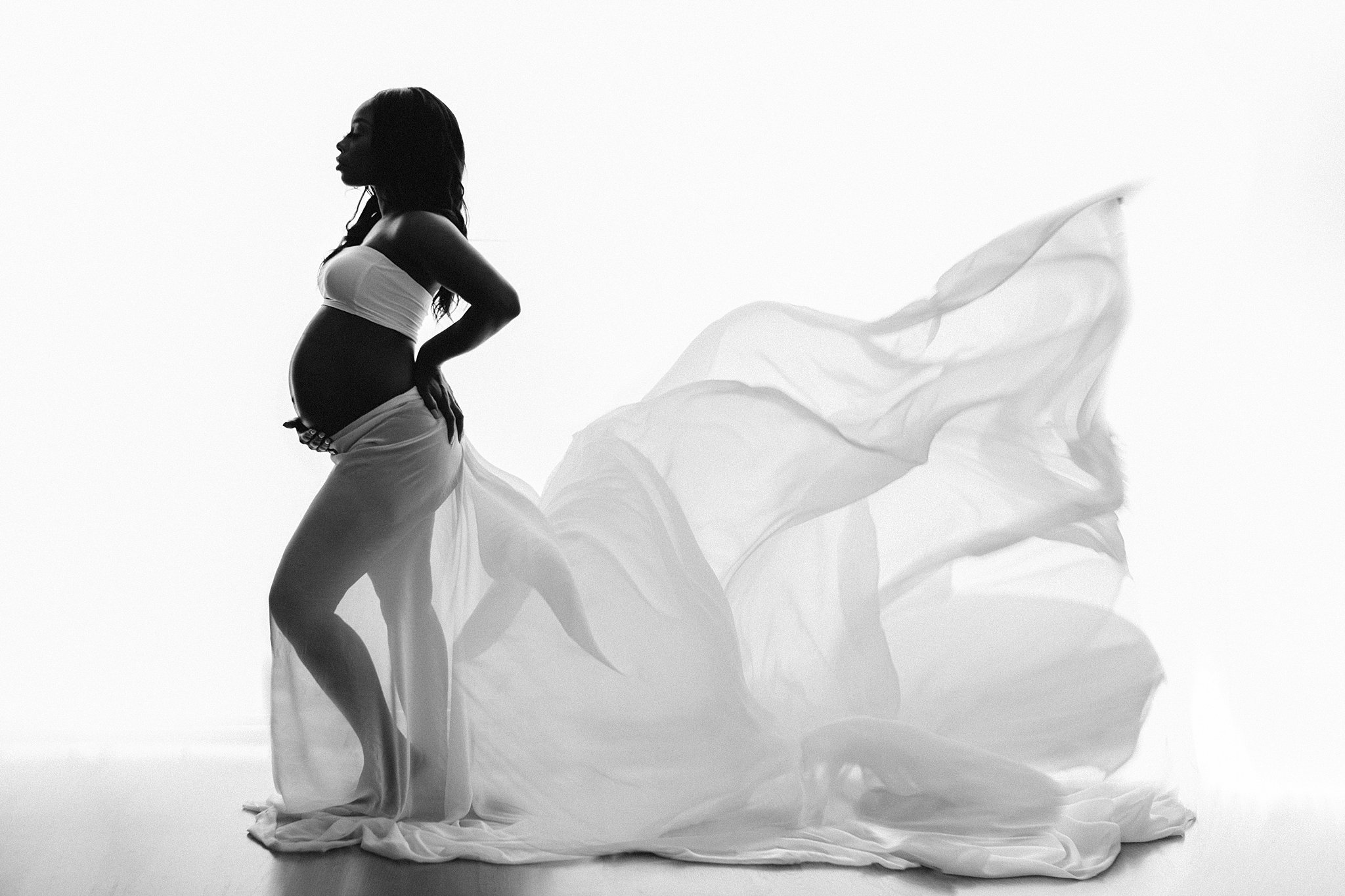 Mother to be stands in a studio holding her back and bump in a long flowing skirt birth by grace midwifery