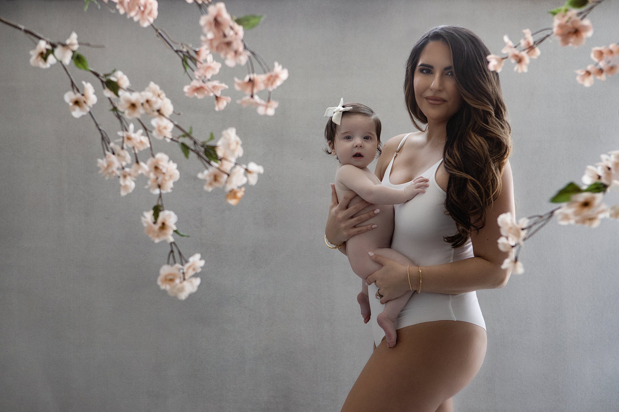 Mother in a white one-piece suit stands holding her daughter in a studio with white flowers