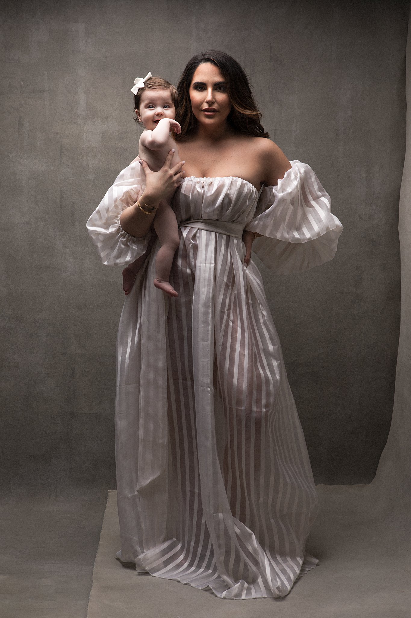 A mother stands in a white dress holding her daughter with a white bow born baby shop
