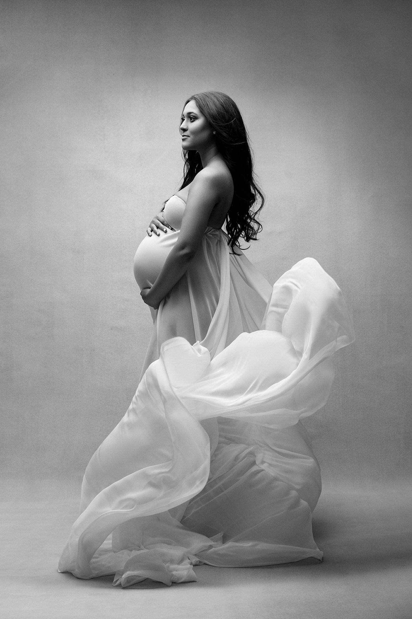 A mother to be holds her bump in a white maternity gown in a studio