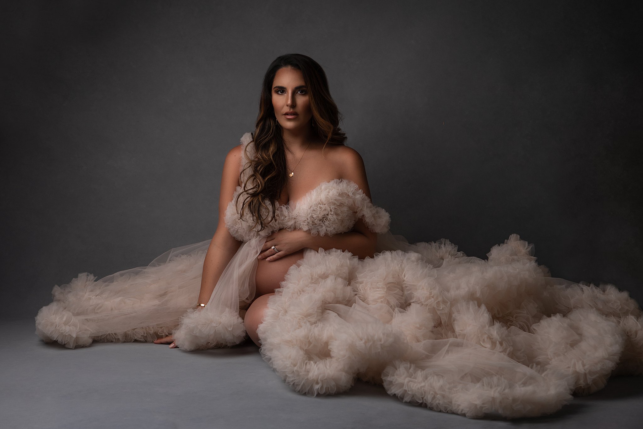 Brown hair mother to be sits on a studio floor wearing a tan velour puff gown maternity stores atlanta
