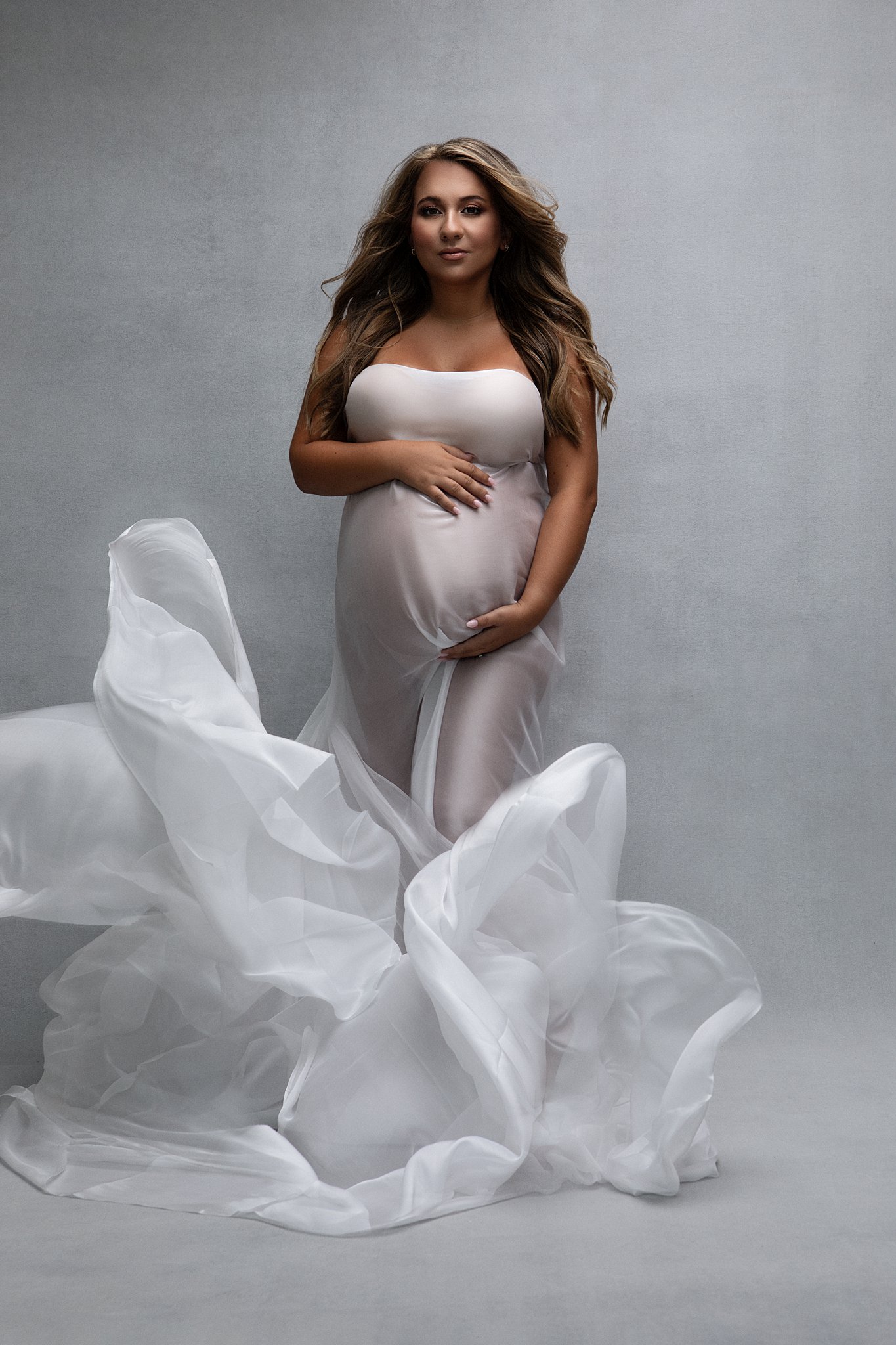 A pregnant woman stands in a studio in a sheer white maternity gown