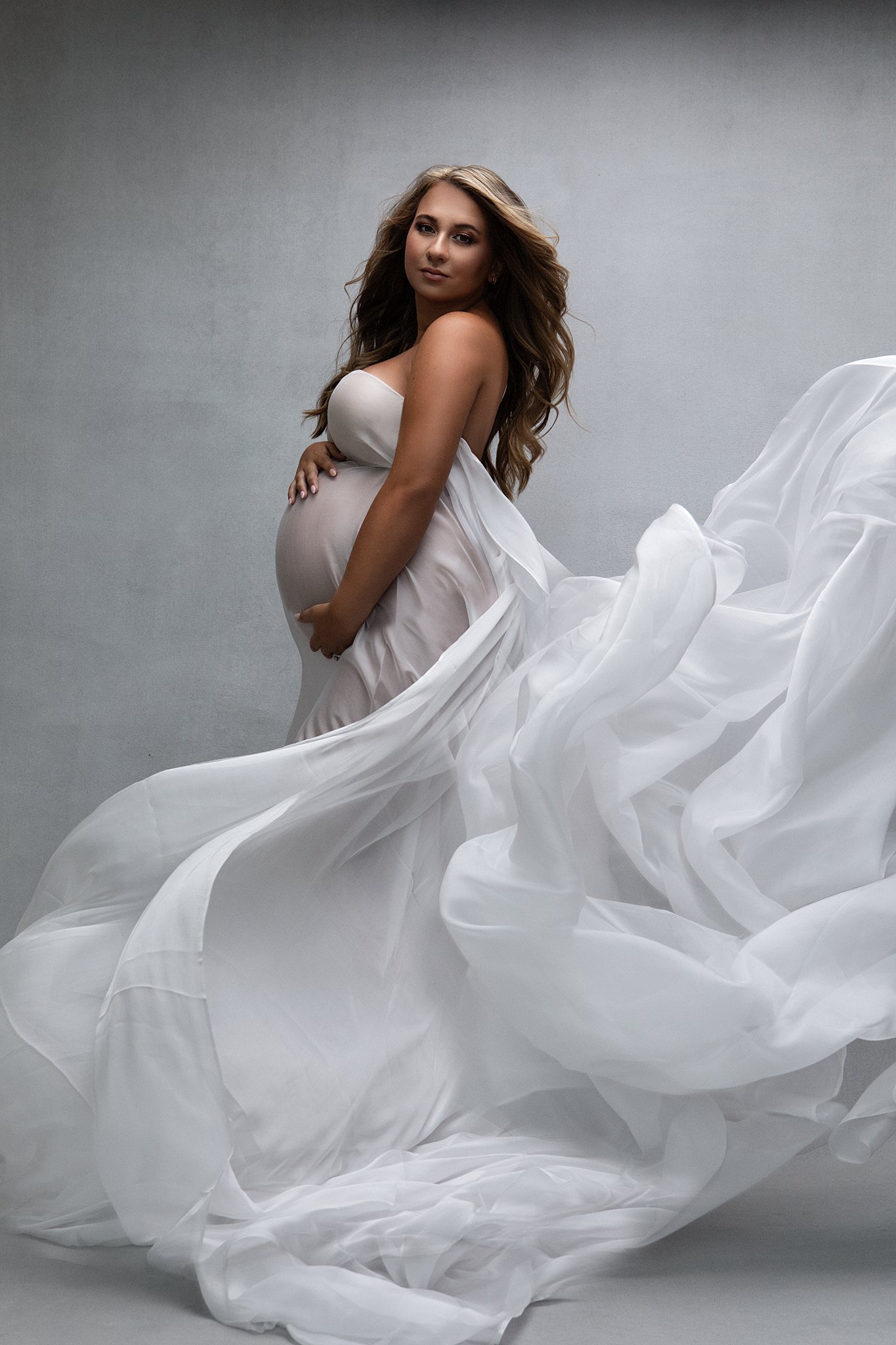 Mother to be stands in a studio holding her bump with fabric flowing through the air around her prenatal yoga plus
