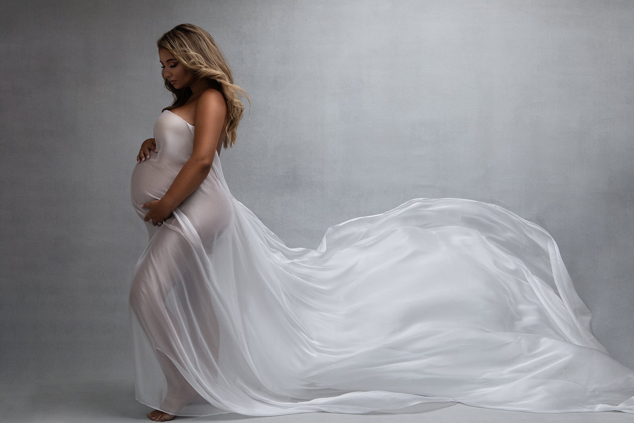 A mom-to-be stands in a studio wearing a sheer maternity gown that is flowing behind her prenatal yoga plus