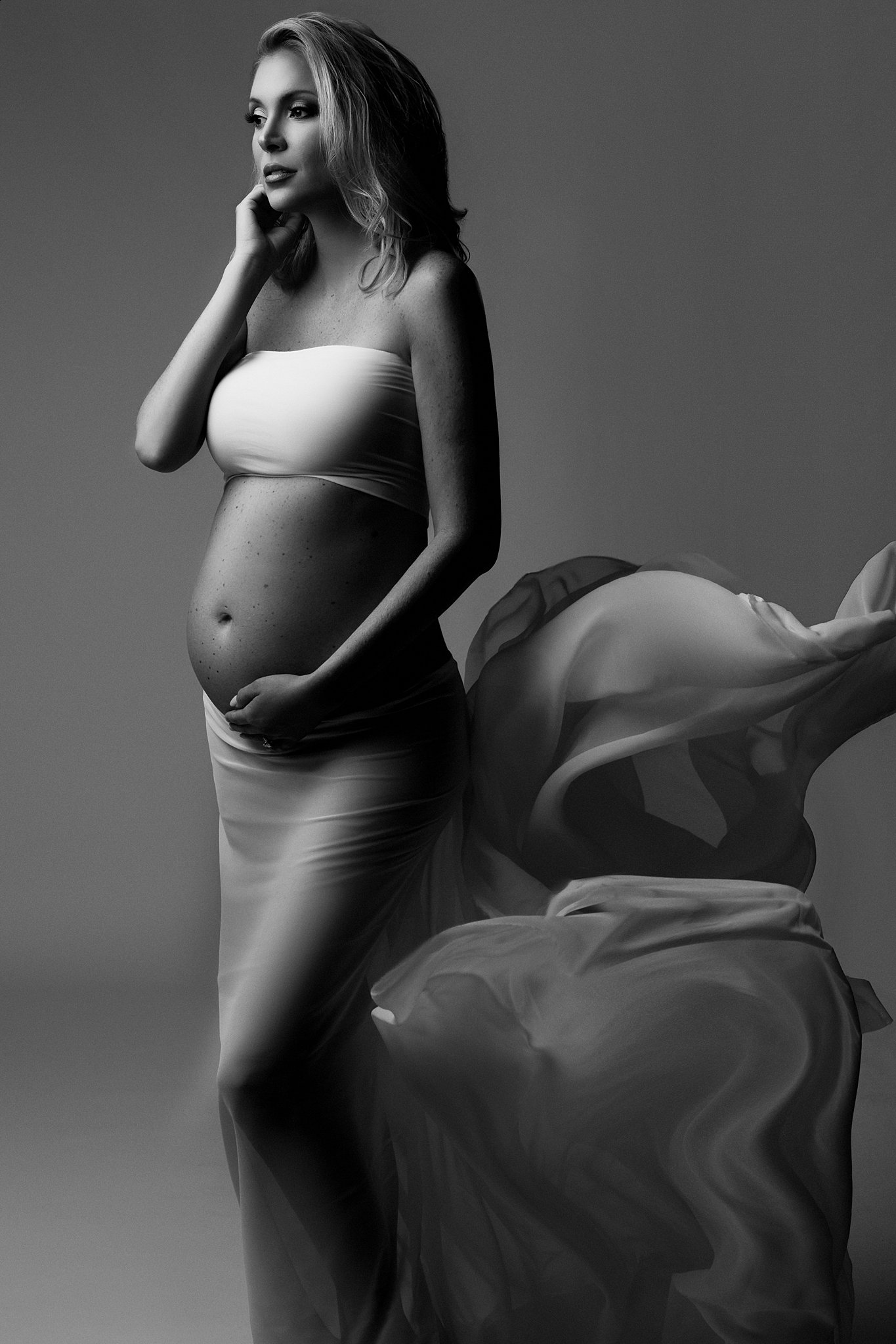 Mother to be wearing a long flowing skirt stands in a studio holding her bump swan coach house atlanta