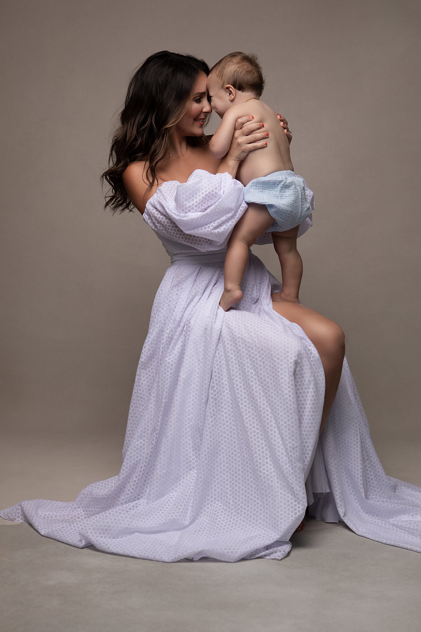A mother in a lilac dress and long brown hair plays forehead to forehead with her toddler in a studio on a stool Mitylene
