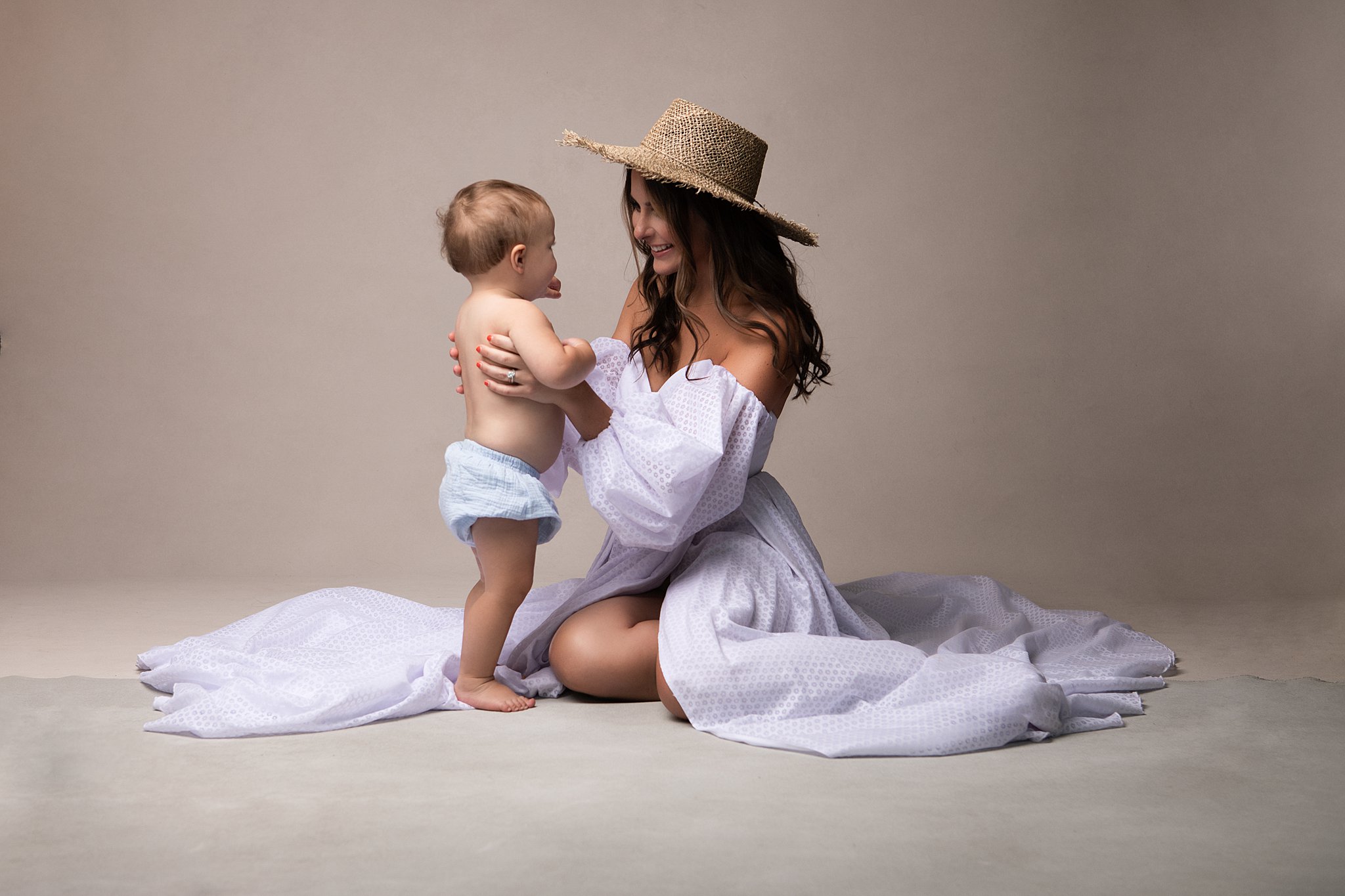 A mother in a straw hat and long flowing lilac dress sits on the floor of a studio playing with her toddler in blue diaper Mitylene