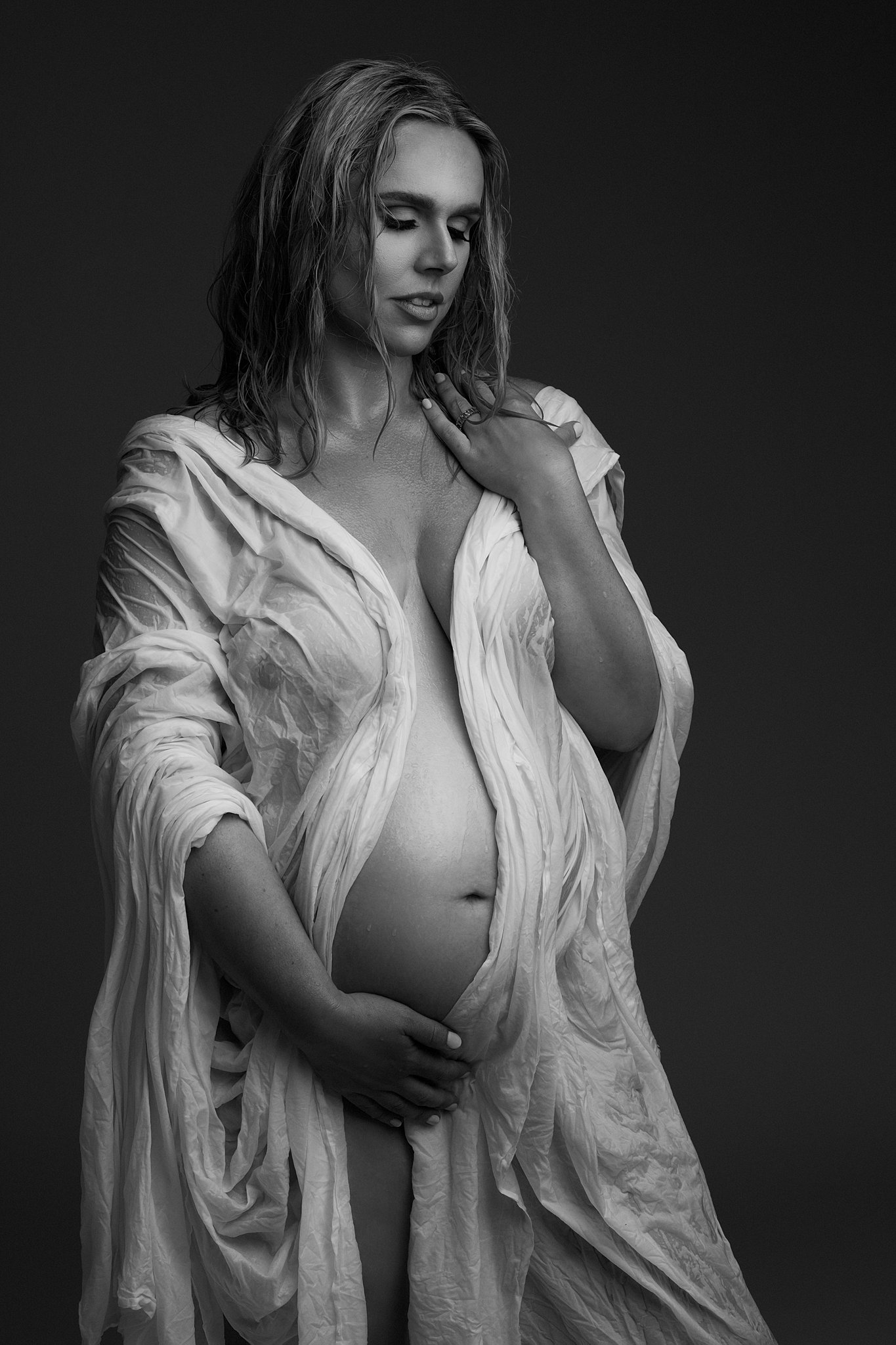 A mom to be draped in a wet white sheet stands in a studio with one hand on her shoulder
