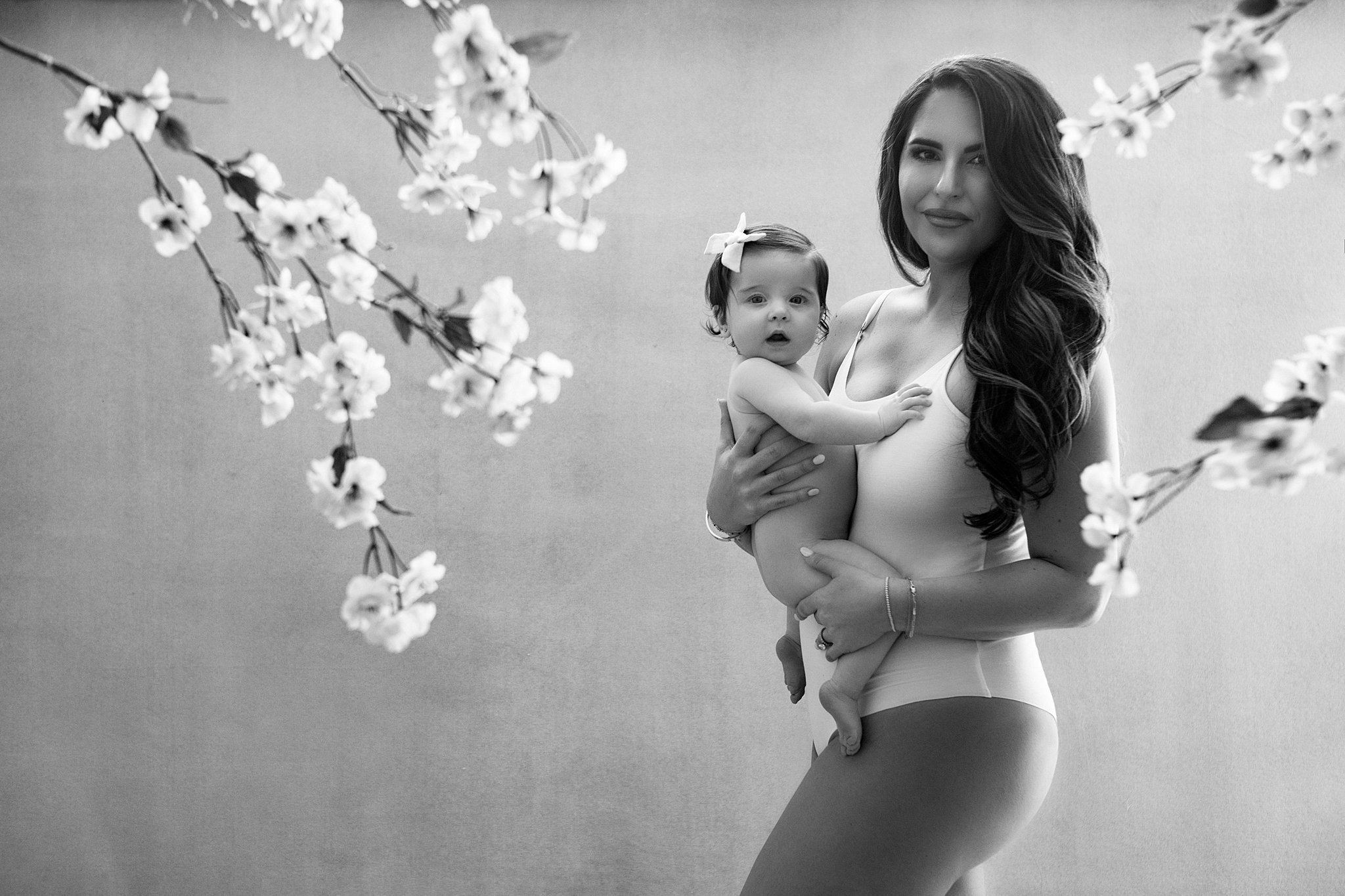 A woman in a white one piece suit holds her toddler baby girl while standing in front of some cherry blossoms in a studio