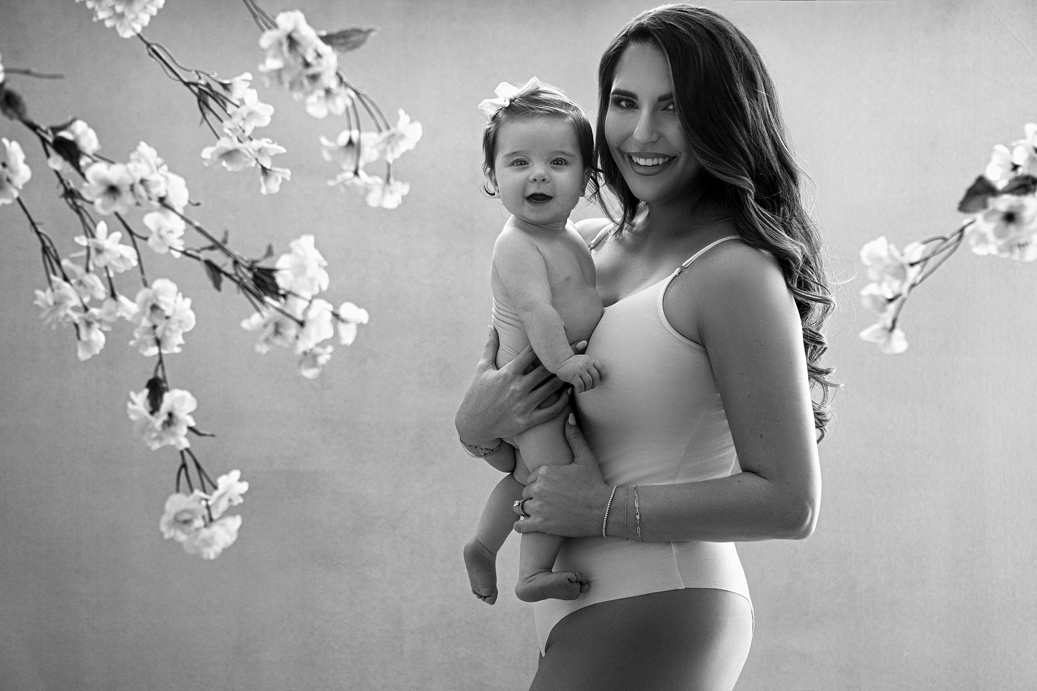 A woman in a white one-piece suit stands amongst some cherry blossoms in a studio while holding her toddler baby