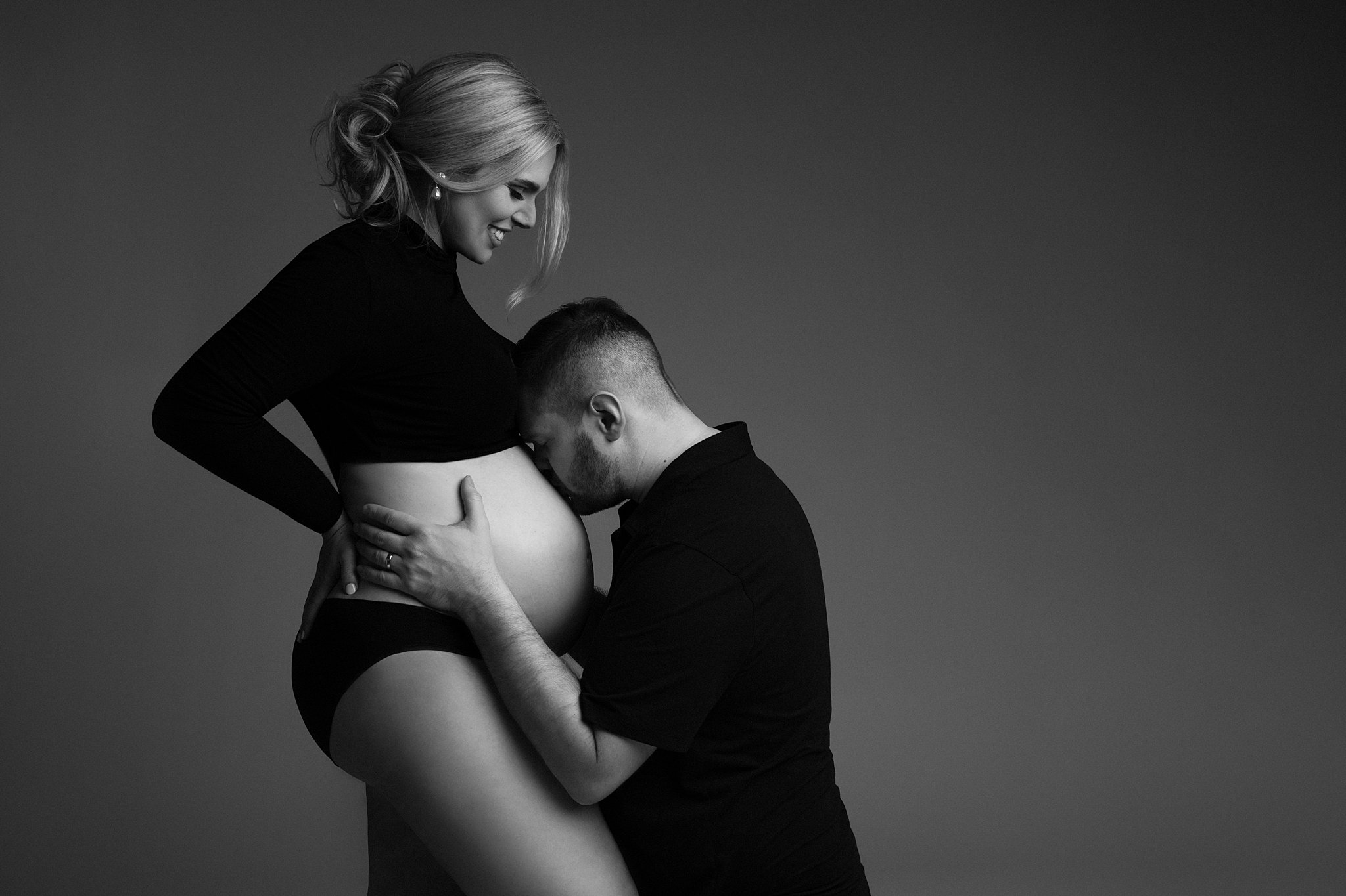 A father on his knees kisses the bump of his pregnant wife while she stands in black underwear and top in a studio bognar & piccolini