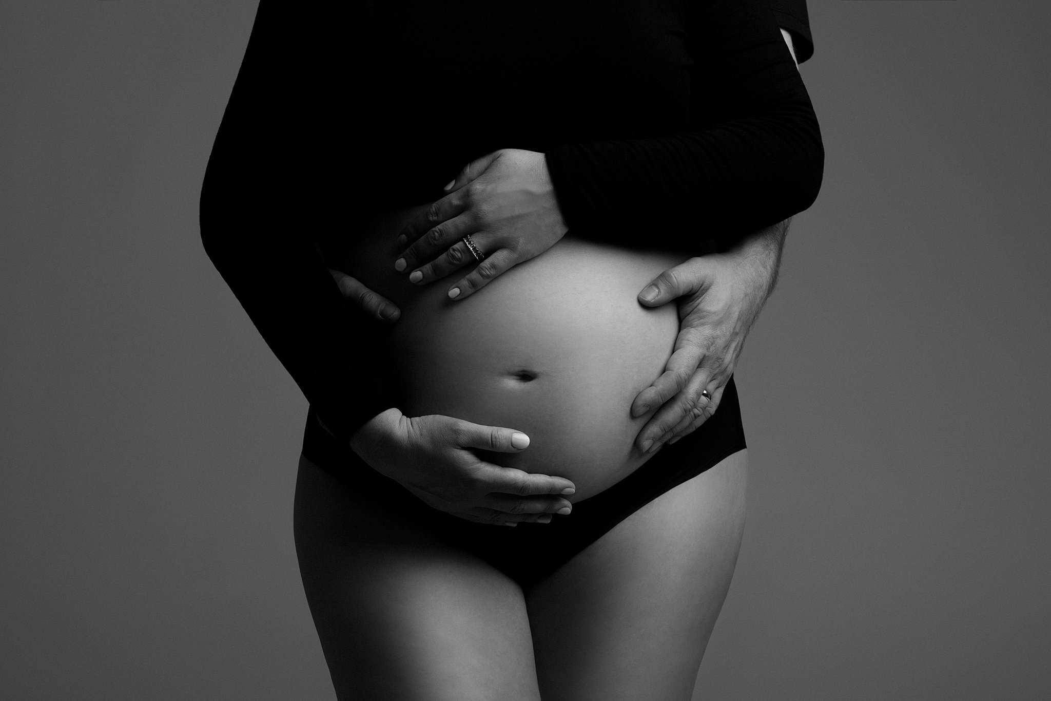 A pregnant woman stands in a studio with her bump exposed with the father reaching from behind to hold it bognar & piccolini