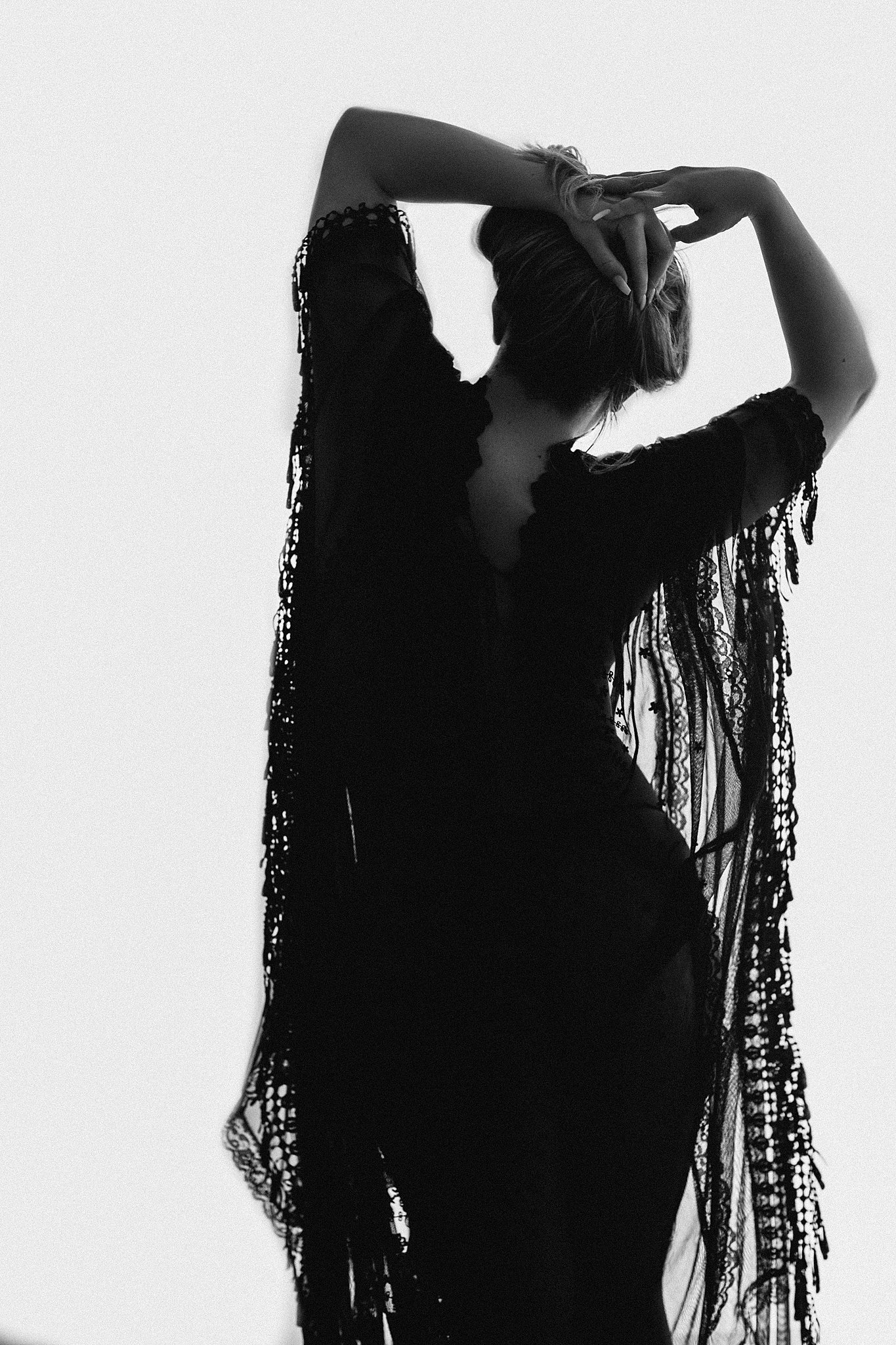 A woman stands in a studio wearing a black shawl with her hands holding her hair up