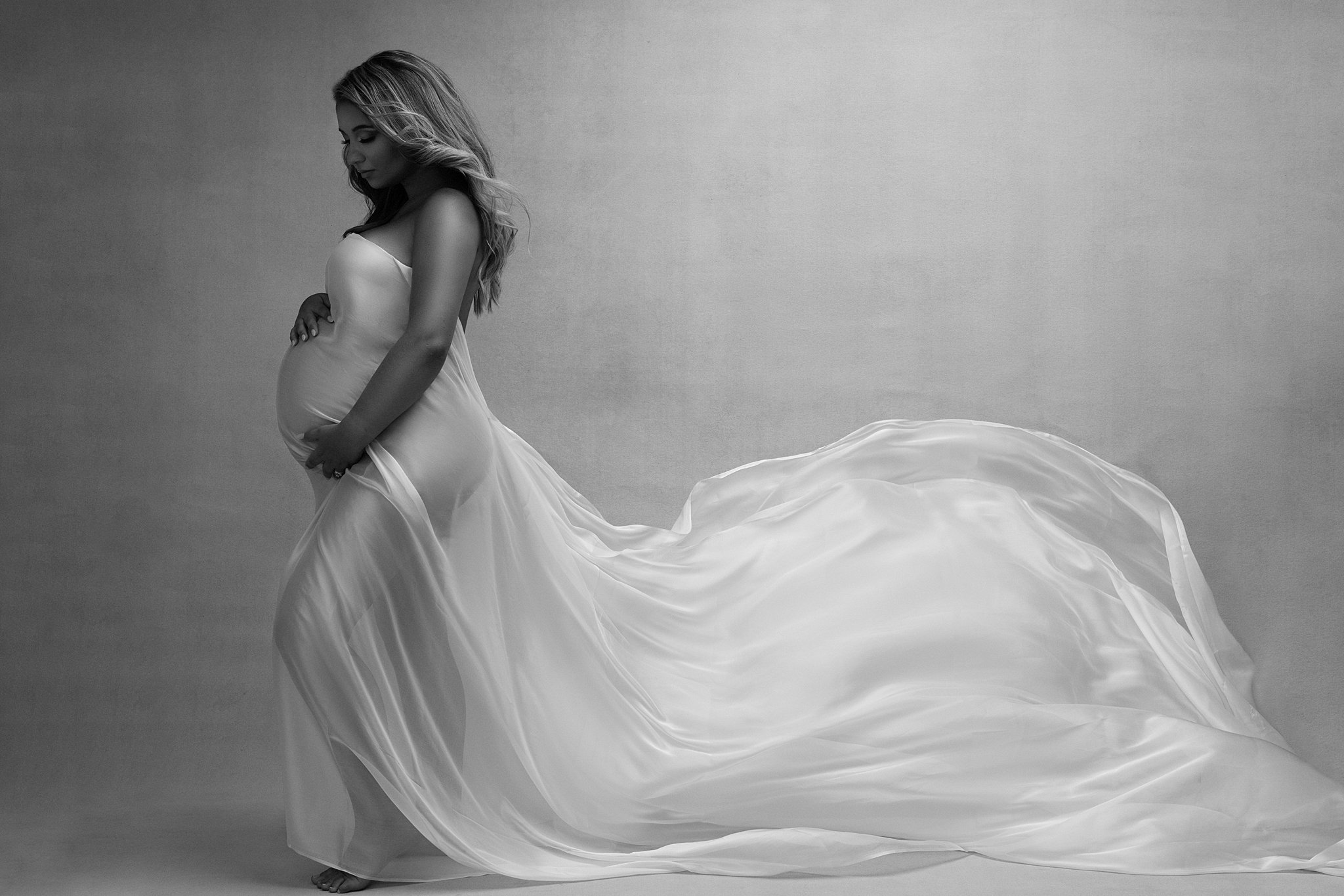 A mom to be stands in a studio wearing a long flowing white maternity gown that is dancing in the wind miami baby shower venue