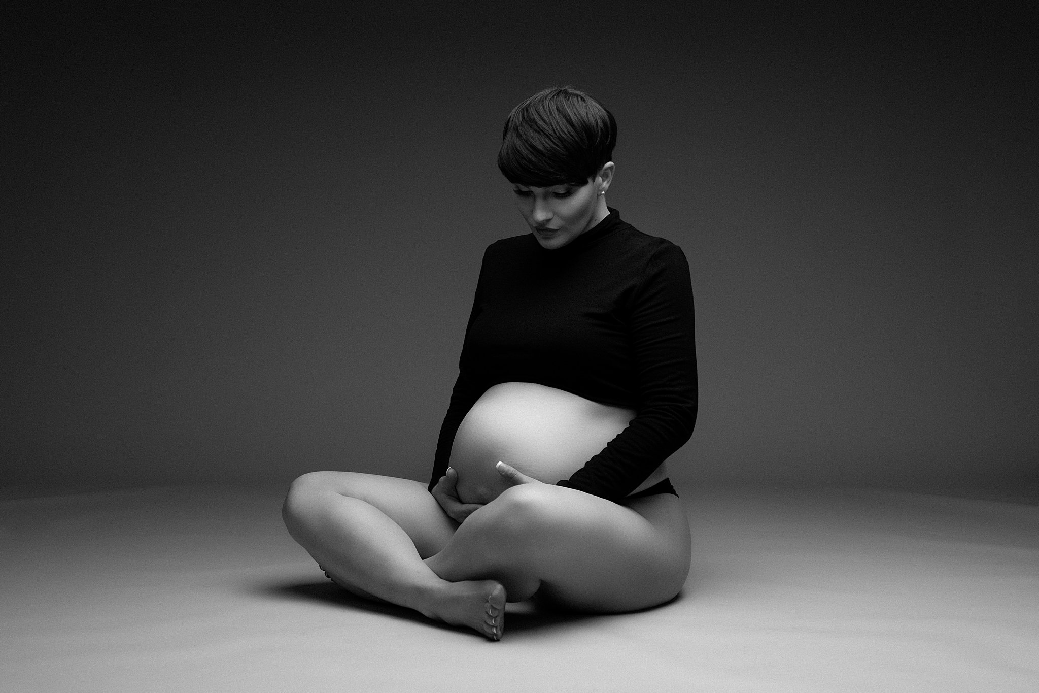 A mother-to-be sits on the floor of a studio cross legged while looking down and holding her bump