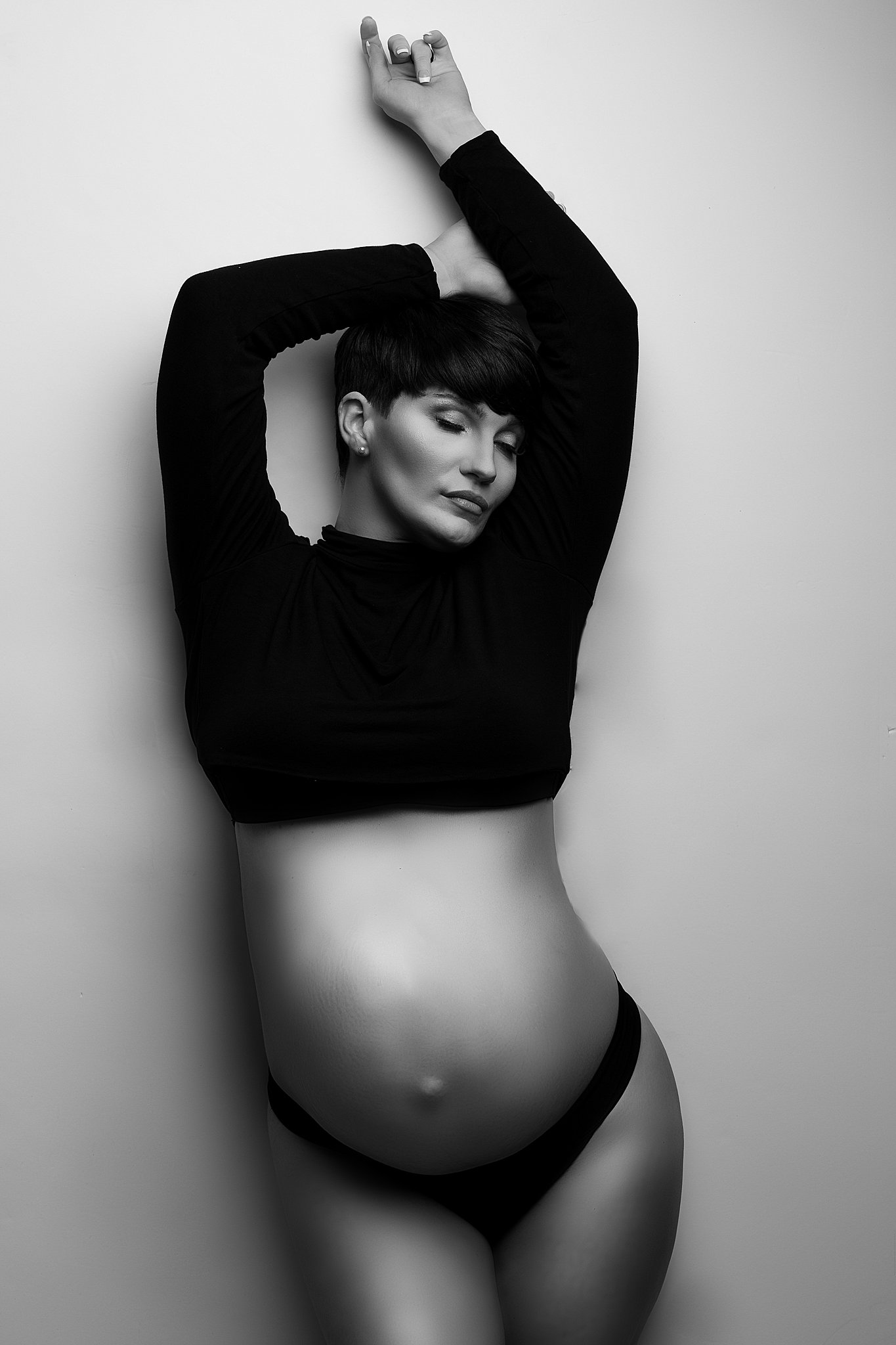A mother to be stands against a white wall wearing a black top and underwear with her belly exposed and hands above her head moonlight midwifery