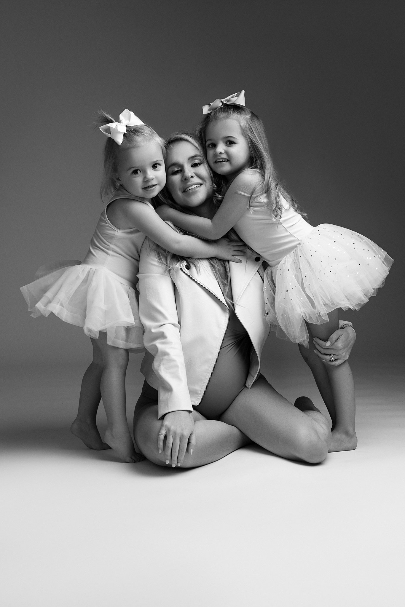 A pregnant mother sits on the floor of a studio while her two daughters hug her while wearing tutus Gootoosh
