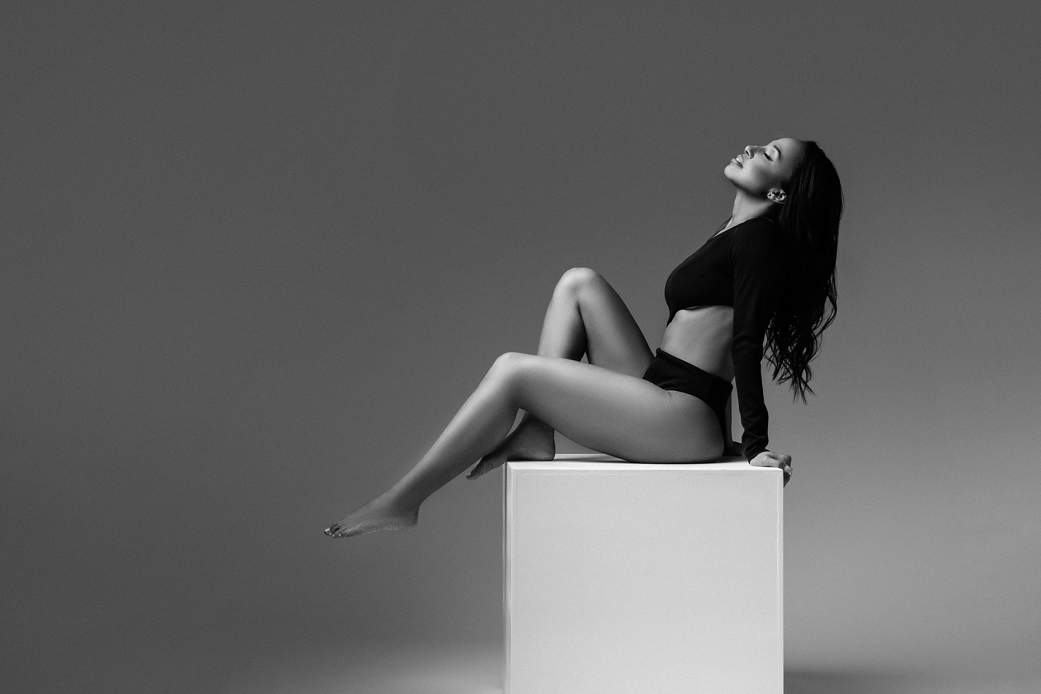 A woman in black lingerie sits on a white cube in a studio