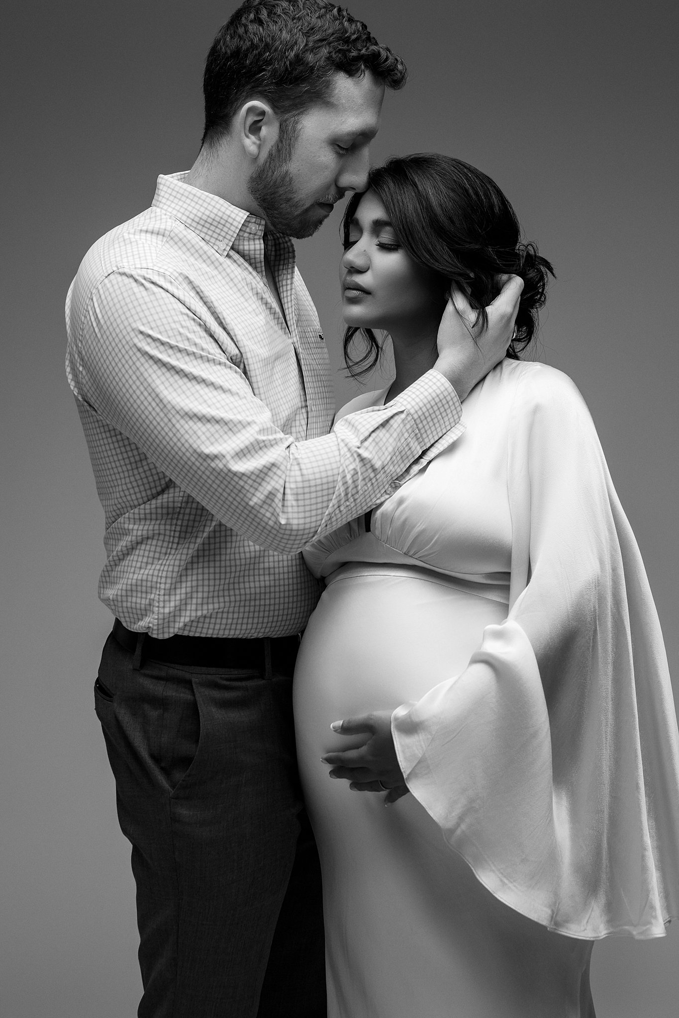 A mother to be in a white maternity dress stands in a studio while her husband leans in to kiss her forehead