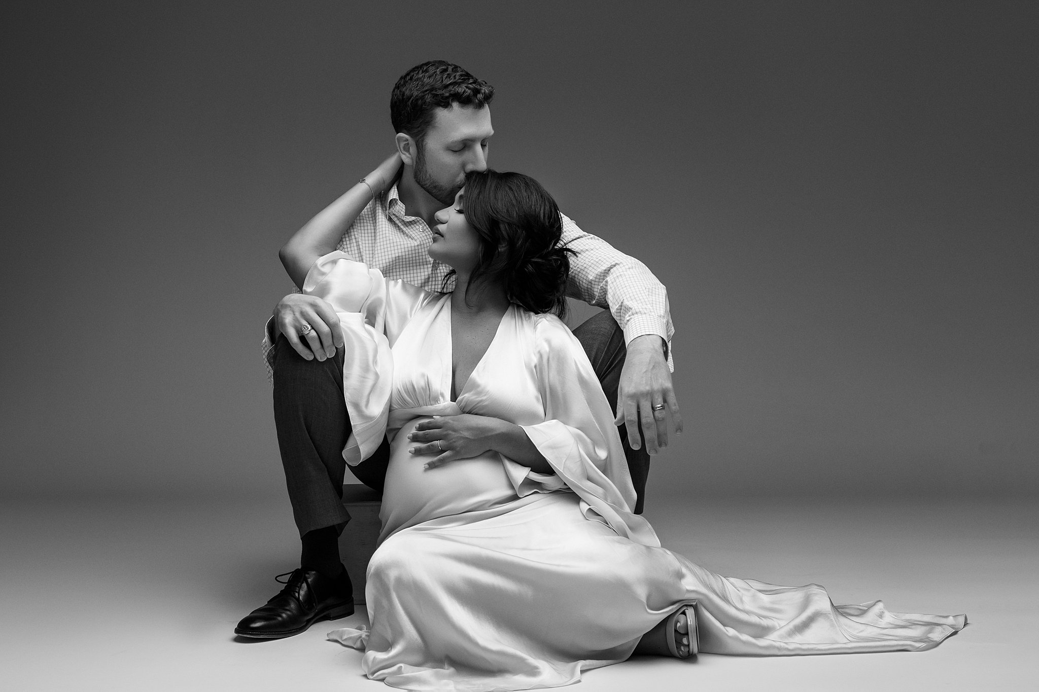 A woman in a white maternity gown sits on the floor of a studio reaching back to her husband behind atlanta toy stores