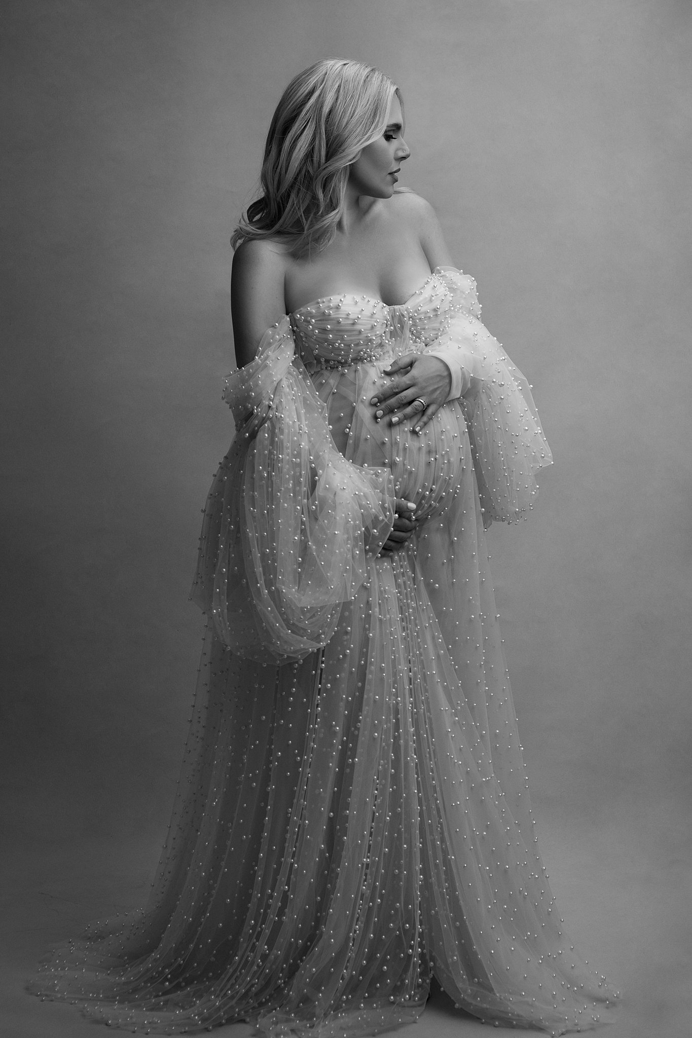 A mom to be in a flowing luxurious maternity gown stands in a studio holding her bump and looking down her shoulder body belly baby
