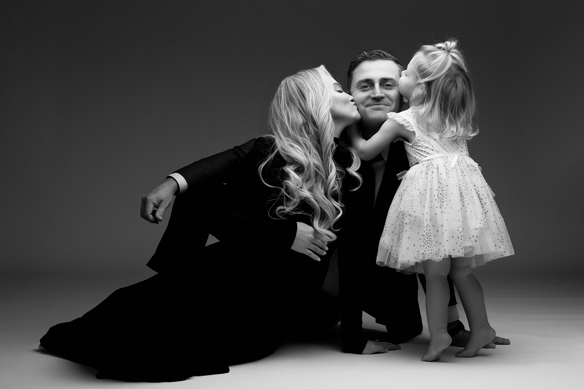 A father sits on the floor of a studio in a black suit while his pregnant wife and toddler daughter kiss his cheeks happy mango