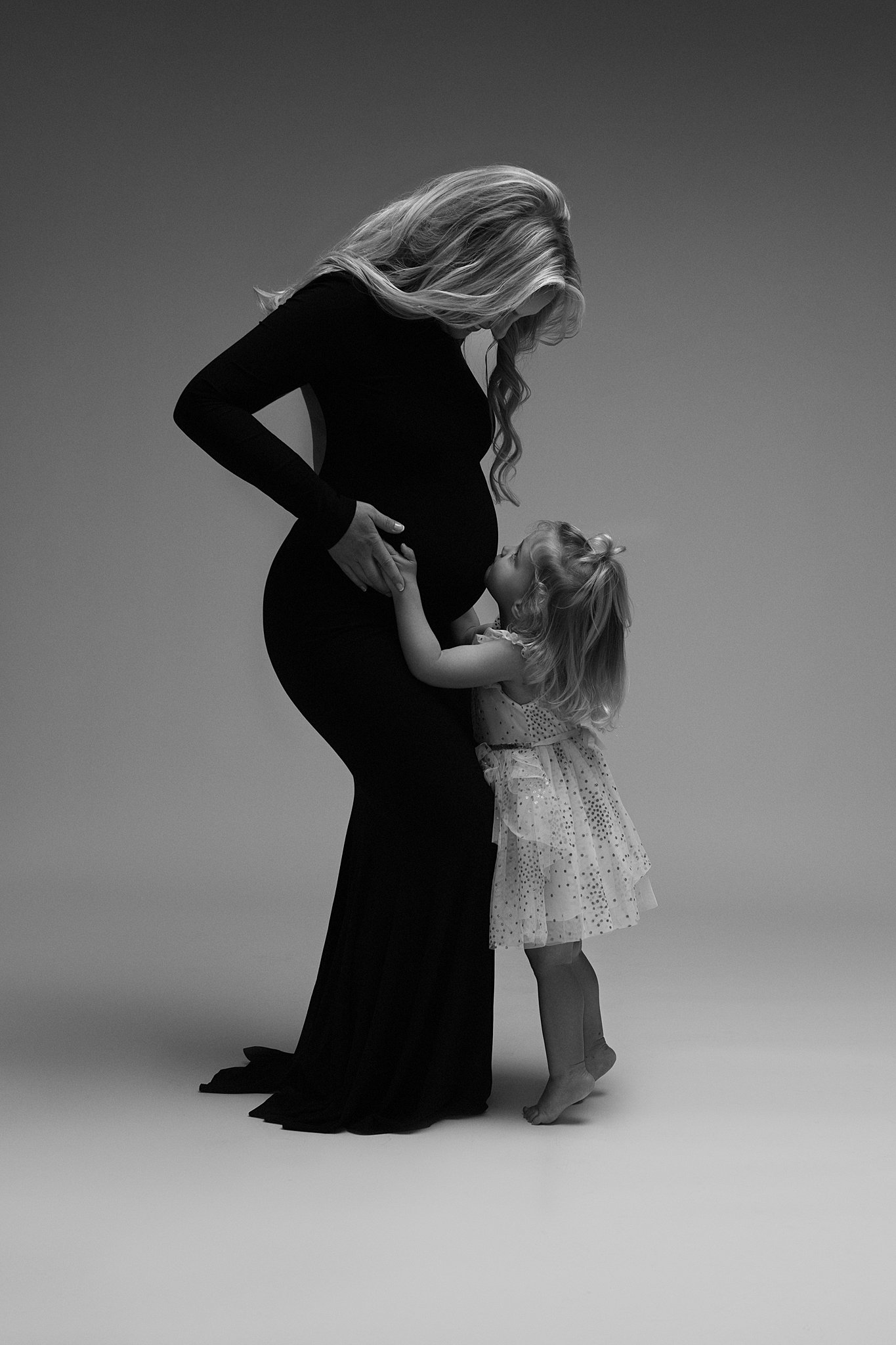 A pregnant woman in a black maternity gown stands in a studio with her young daughter kissing her bump