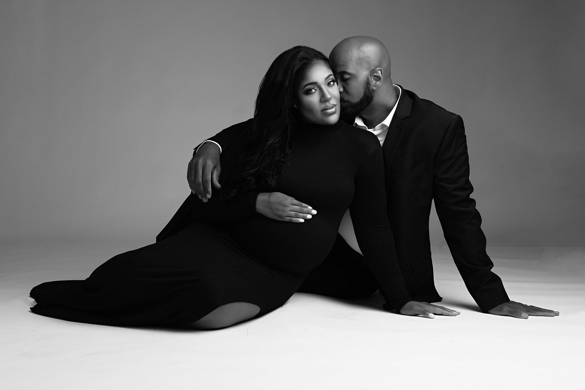 A mom-to-be sits on the floor of a studio, leaning into her husband in a black maternity dress marietta pediatricians