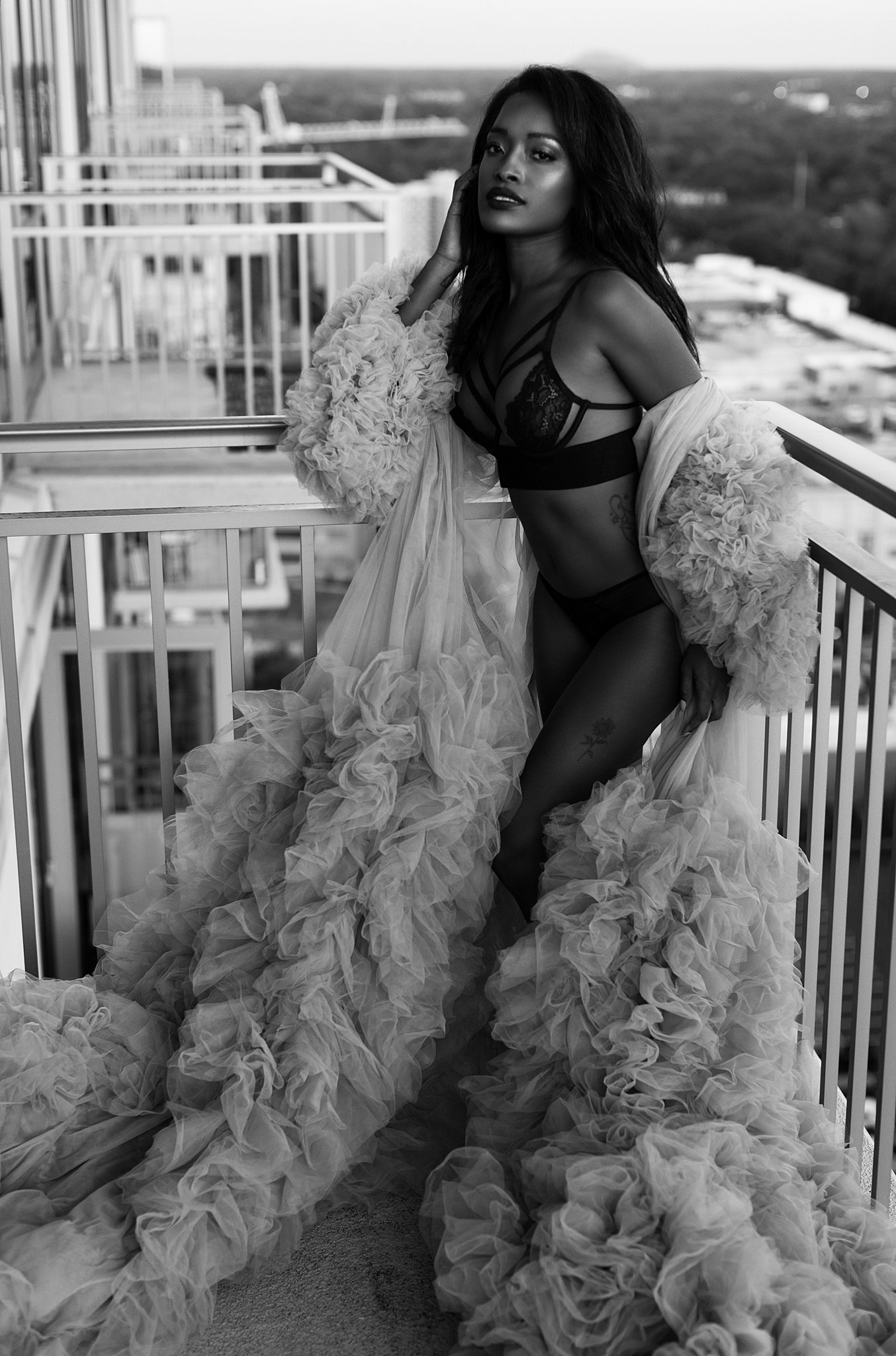 A woman in black lingerie and a tule coverup leans on the railing of a balcony massageluxe miami