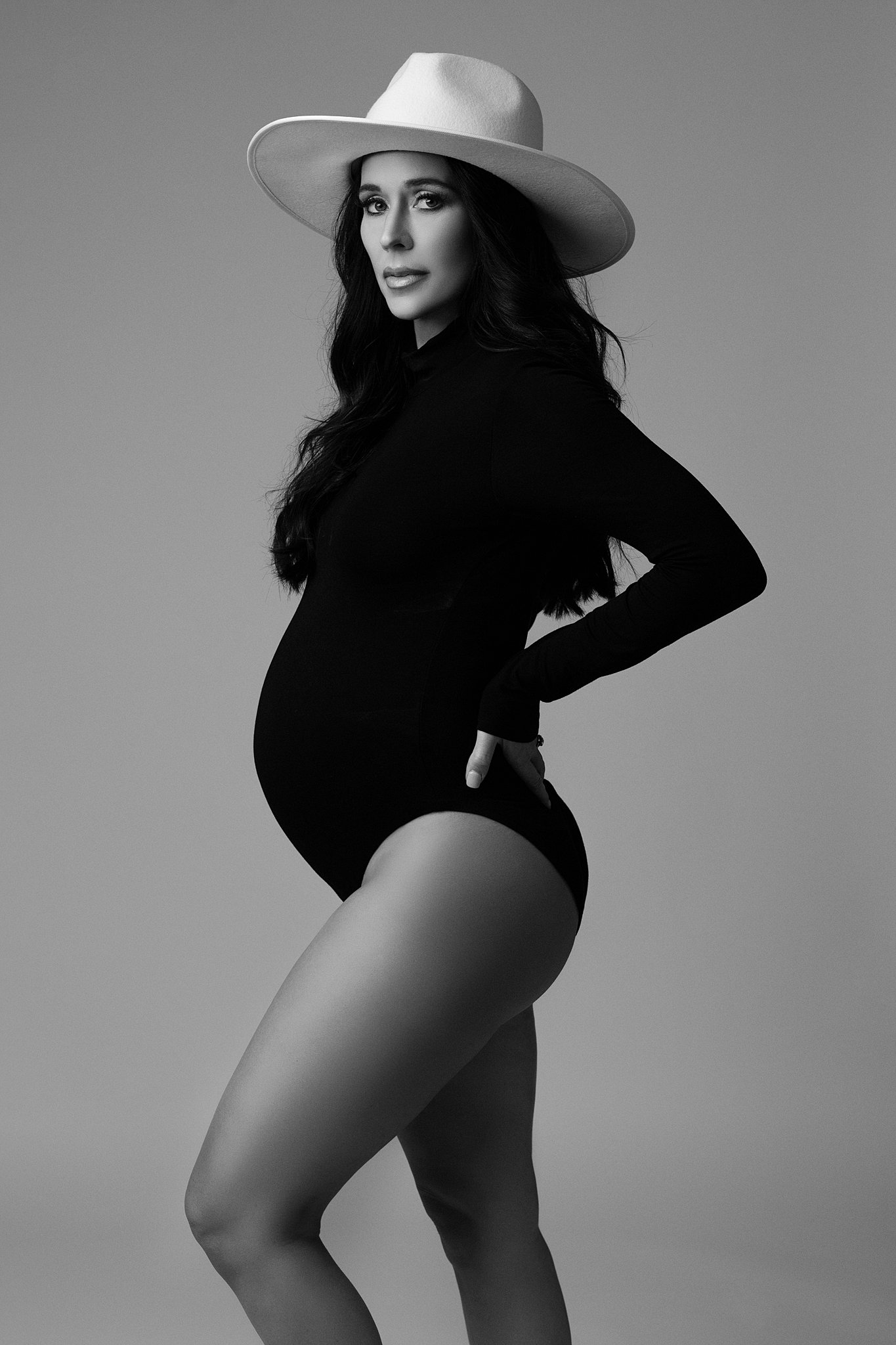 A pregnant woman stands in a studio with a hand on her back in a black one-piece suit sugarboo atlanta