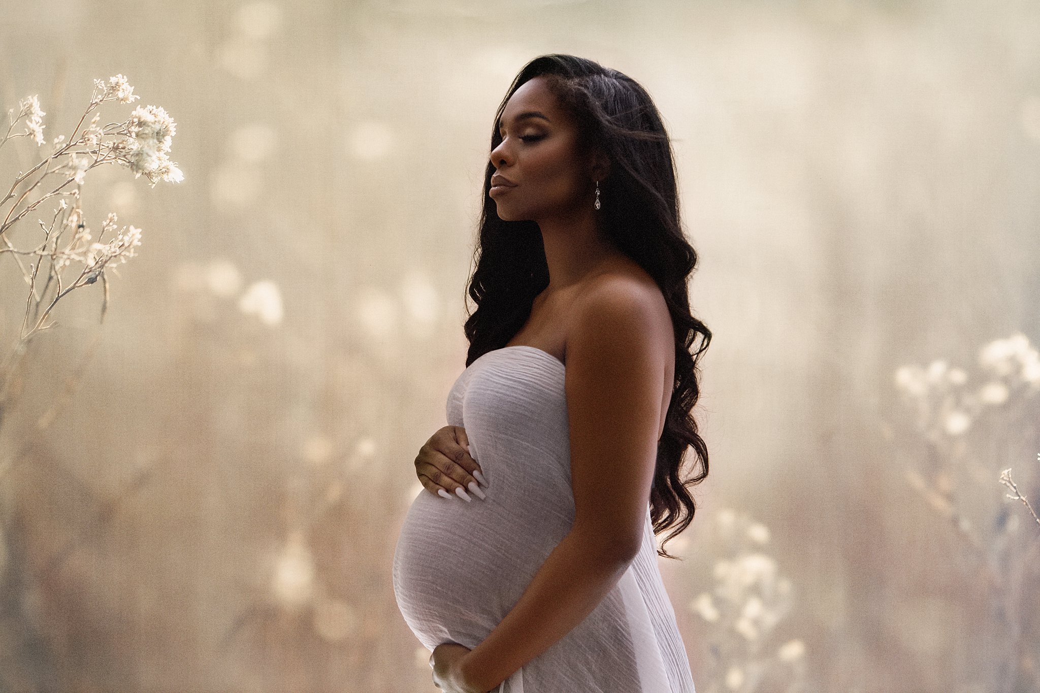 A mother to be stands in a white maternity gown in a studio
