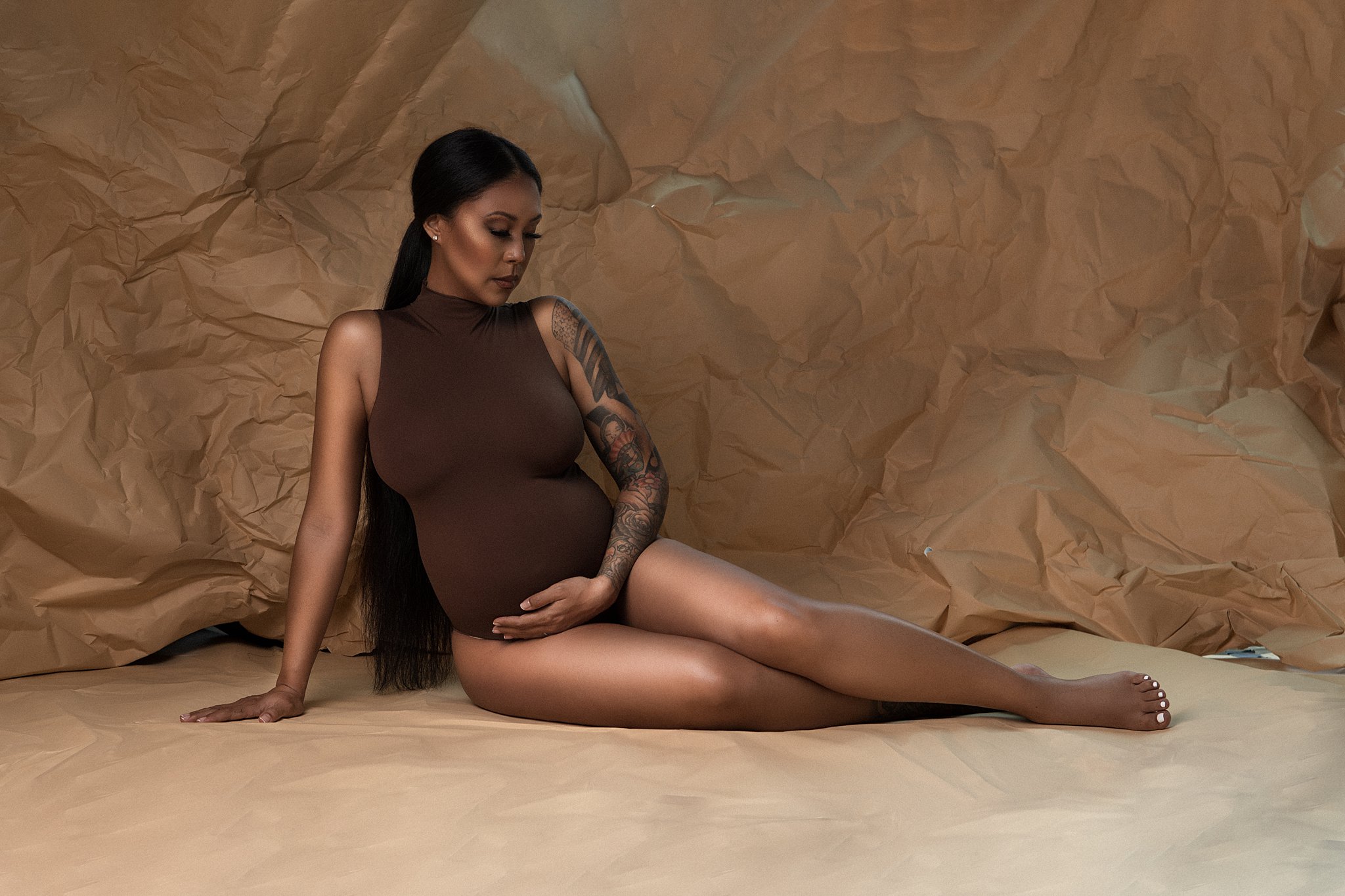 A mother to be in a brown maternity onesie lays on the floor of a studio