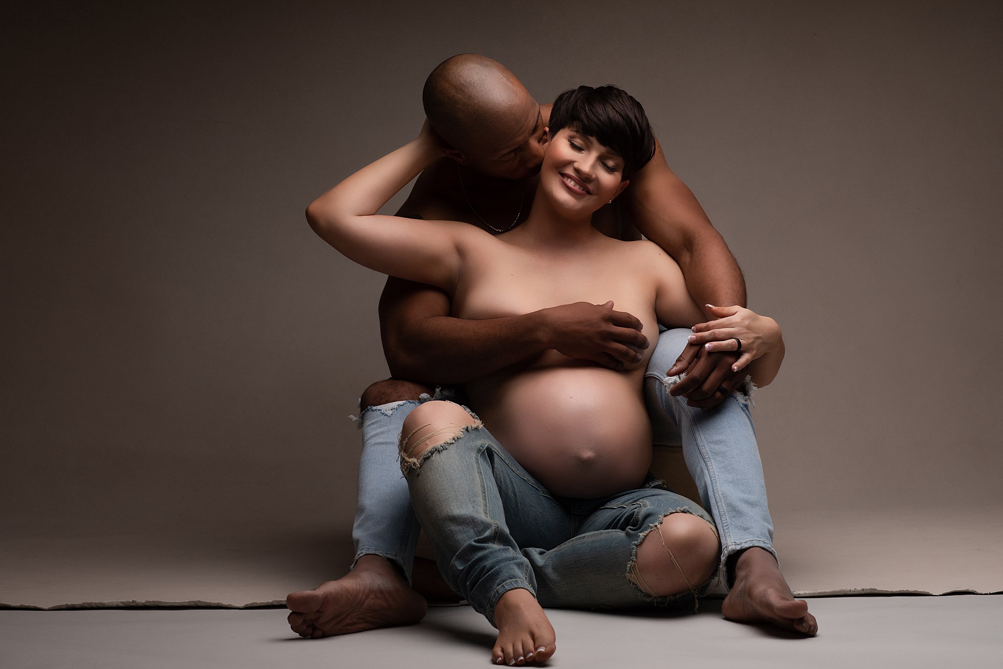 A mom-to-be sits on the floor of a studio leaning back into a kiss from her husband