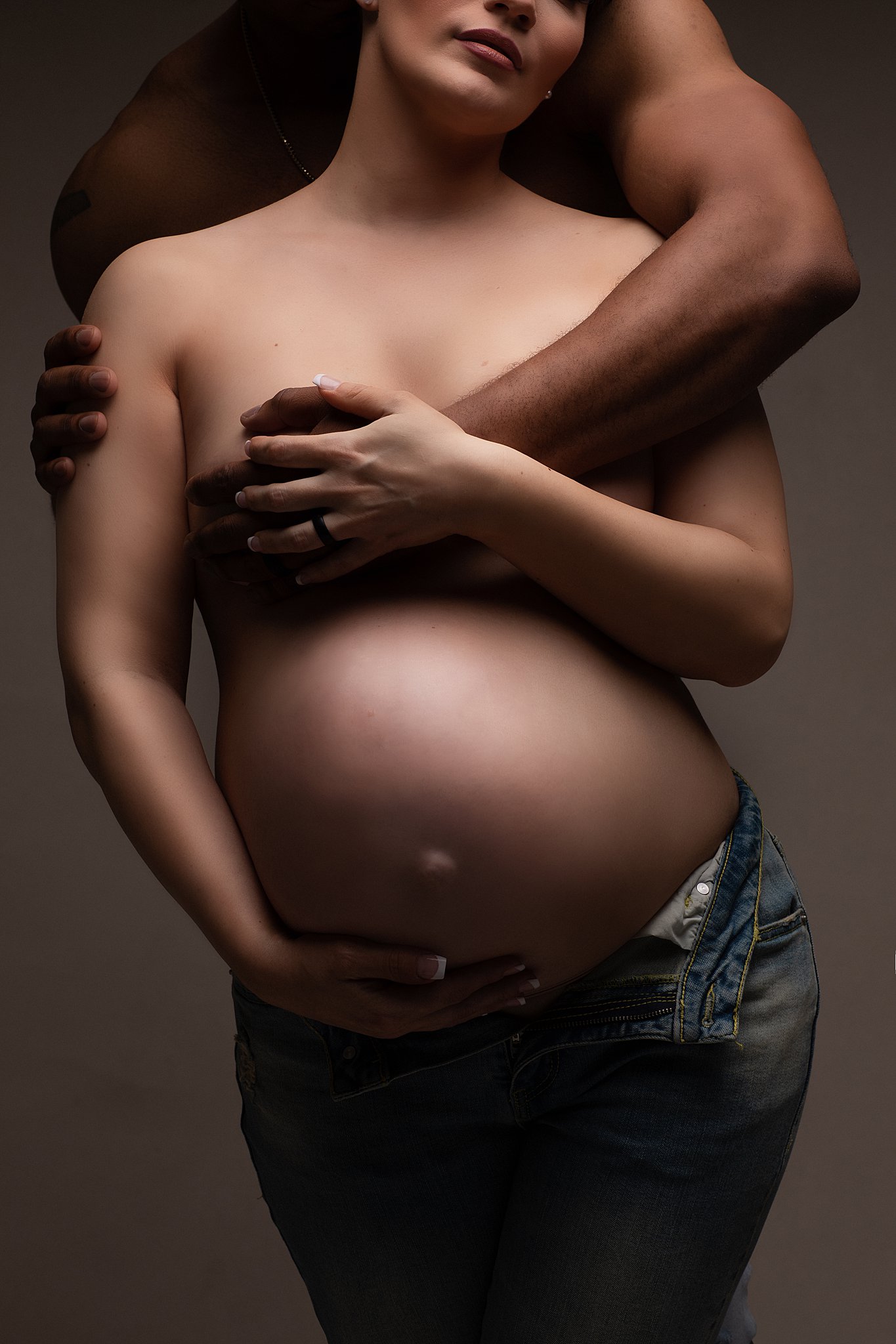 A mother to be holds her bump while standing in a studio being hugged by her husband from behind athens obgyn