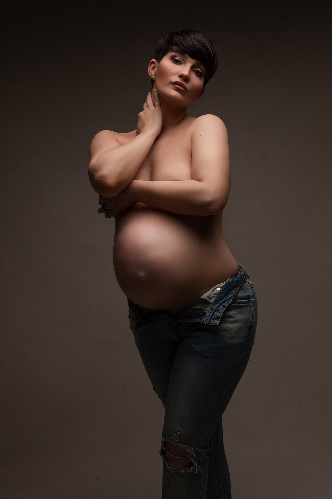 A mother to be stands in a studio in open jeans covering her breasts with her arm athens obgyn