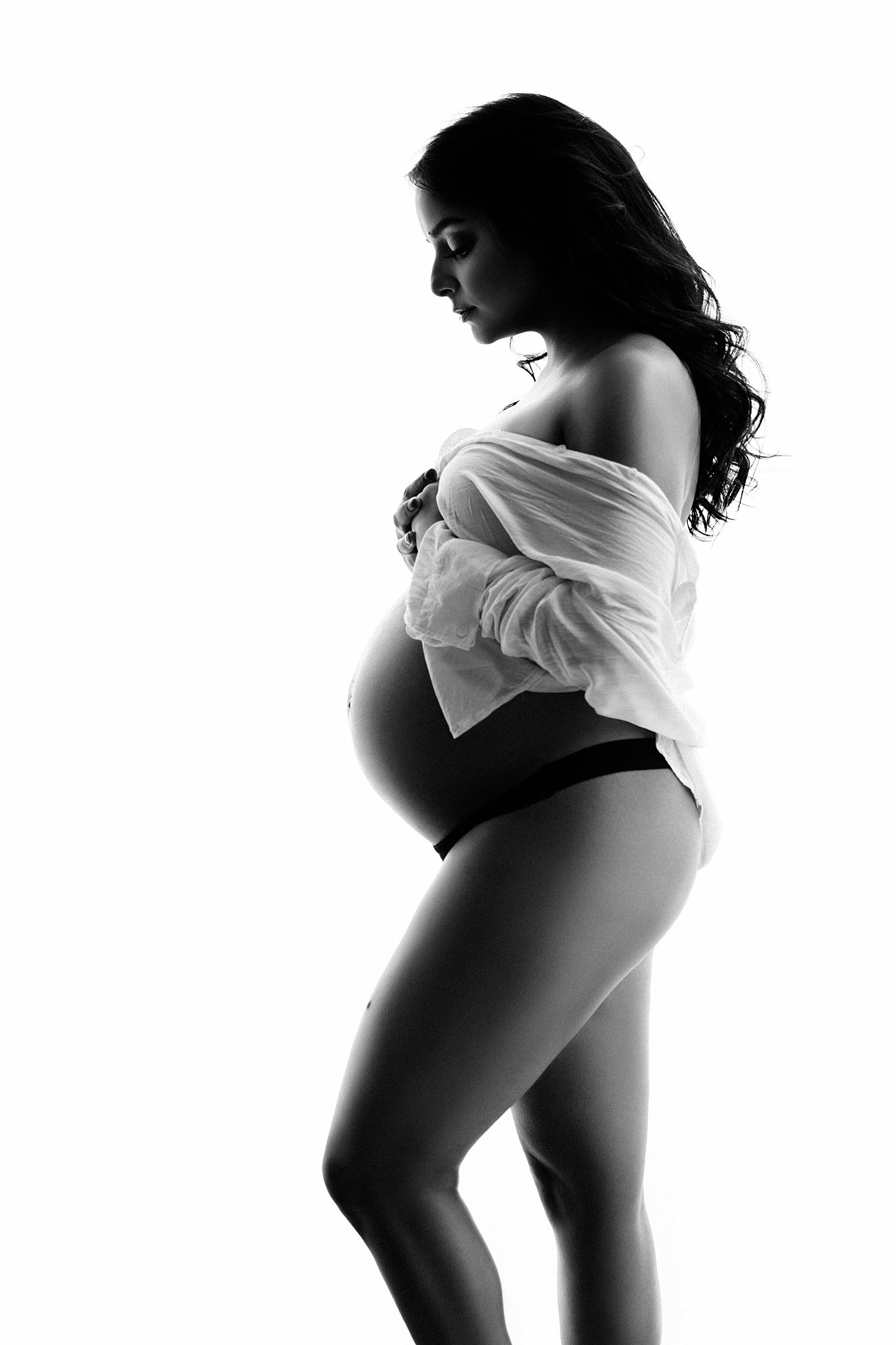 A mother to be wears a white button down shirt and black underwear in a studio looking down at her bump