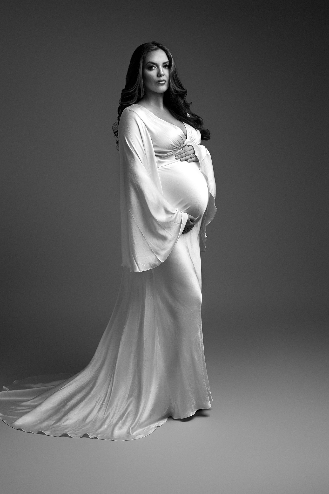 A mom to be stands in a studio holding her bump in a long flowing maternity gown