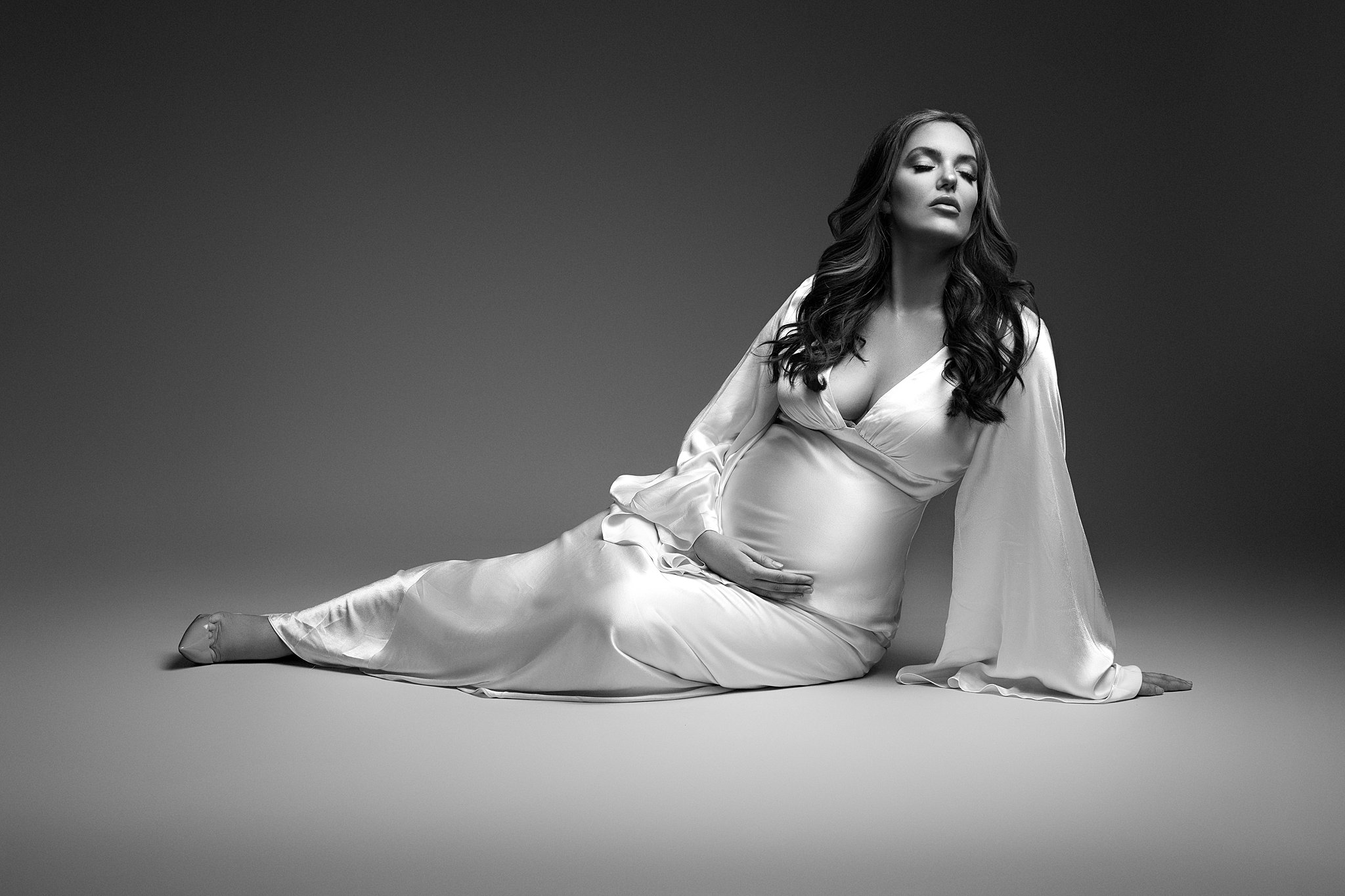 A mom to be lays across a studio floor in a shiny maternity gown holding her bump maternal fetal medicine miami