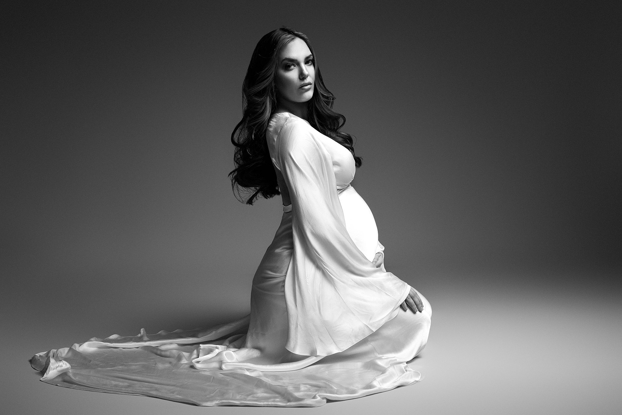 A mother to be kneels on a studio floor in a long white maternity gown maternal fetal medicine miami